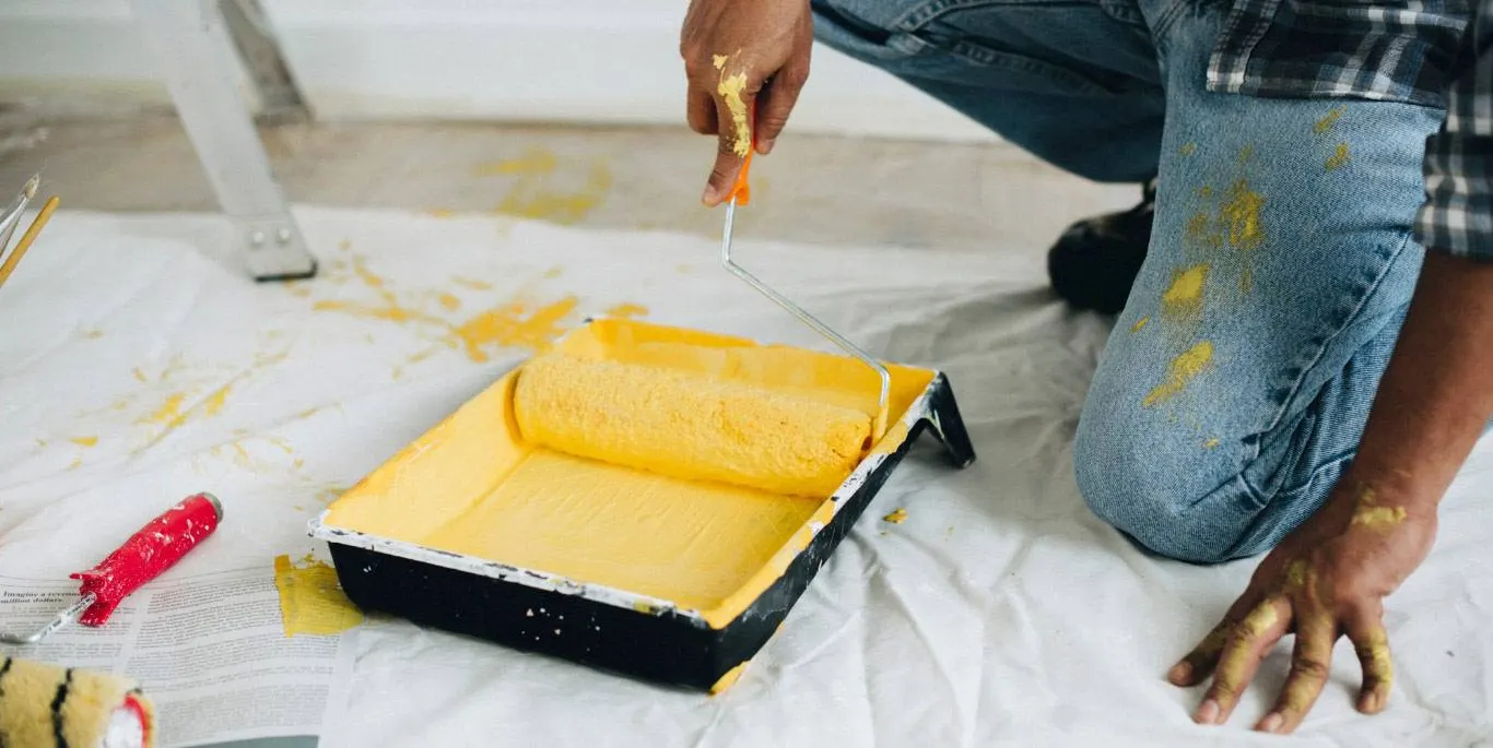 Pintor cargando un rodillo en una bandeja de pintura amarilla sobre un suelo protegido